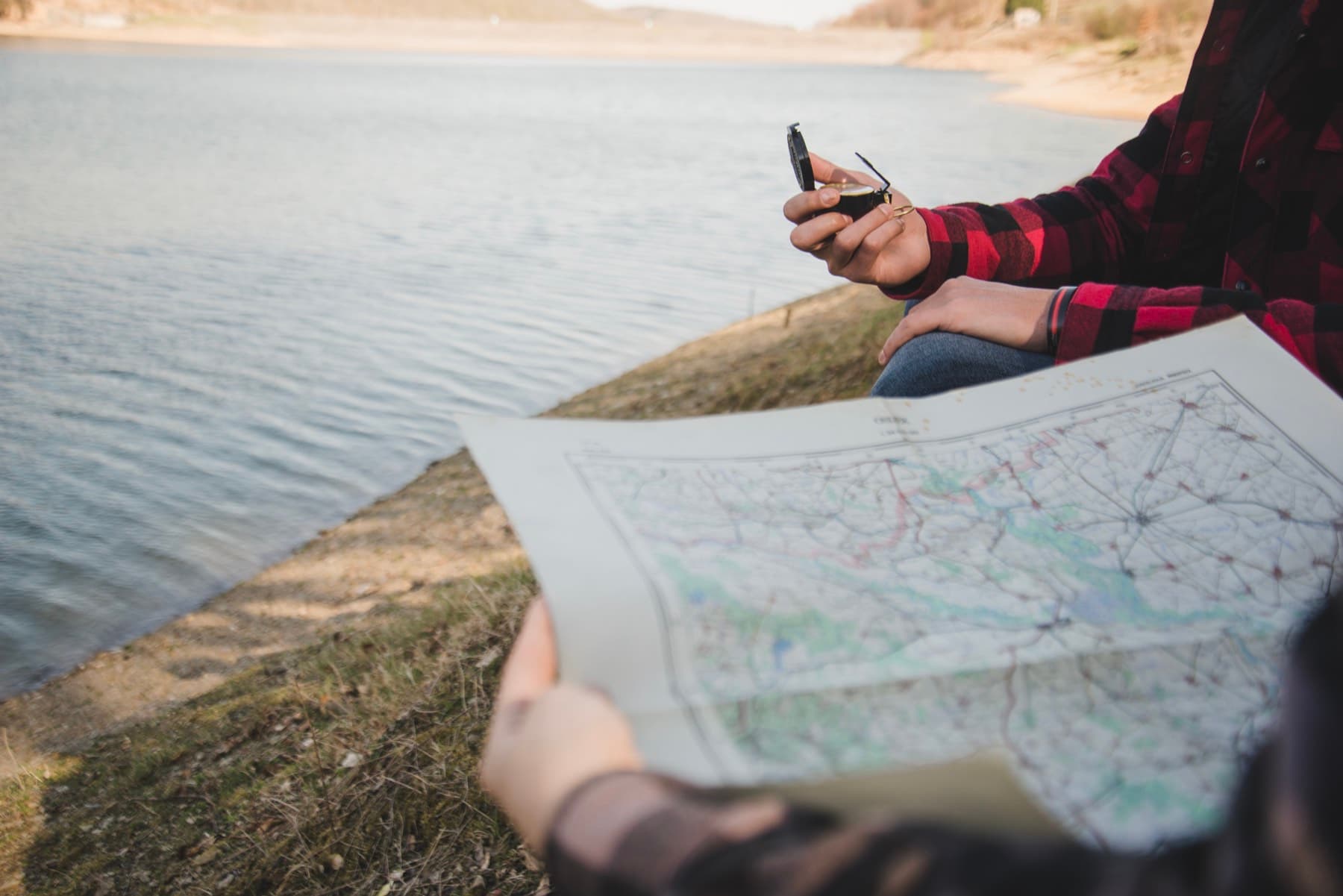 Person seated at the edge of a body of water with a map spread out and a handheld instrument