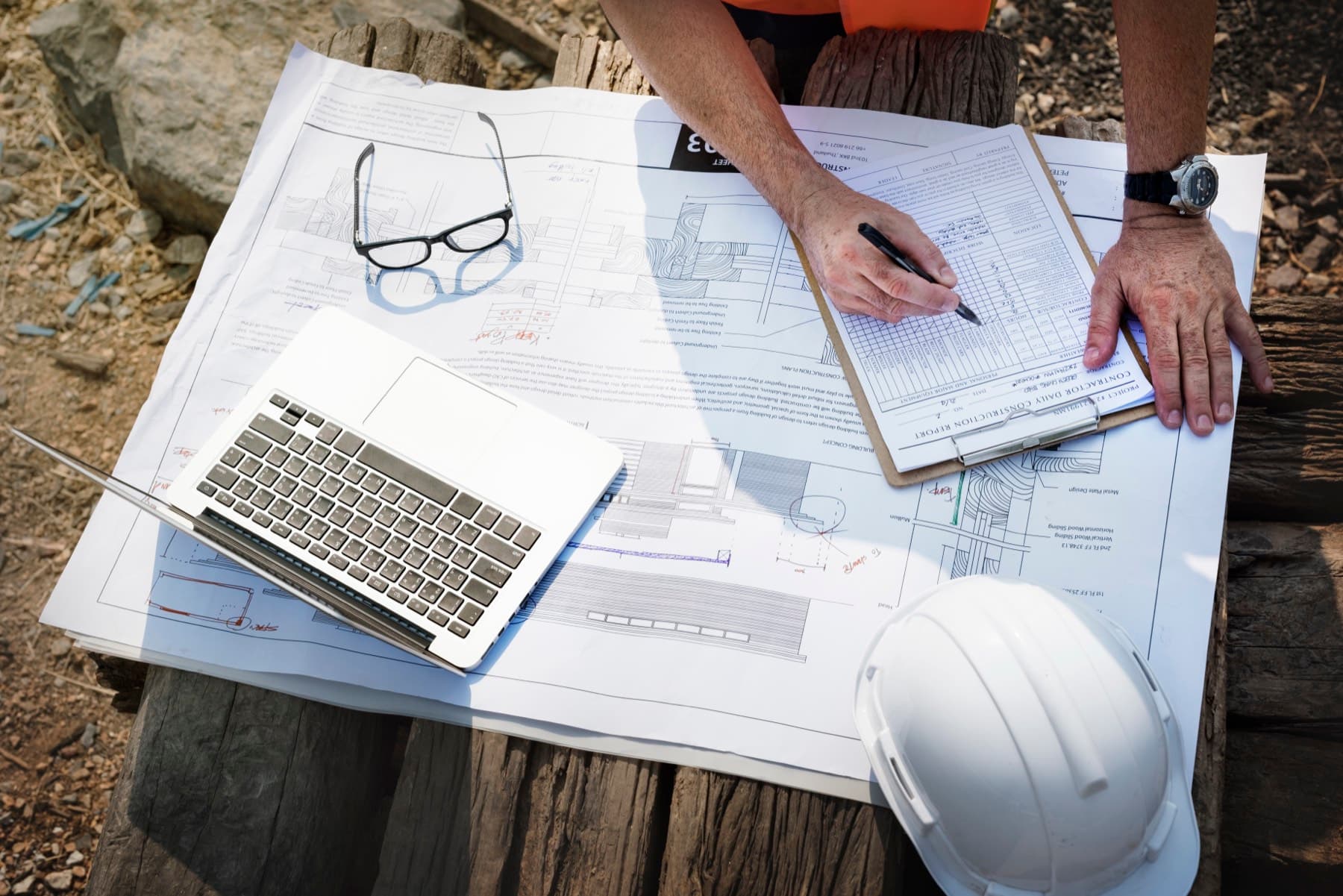 Hands marking up a checklist over construction drawings, beside a laptop, a hard hat and reading glasses