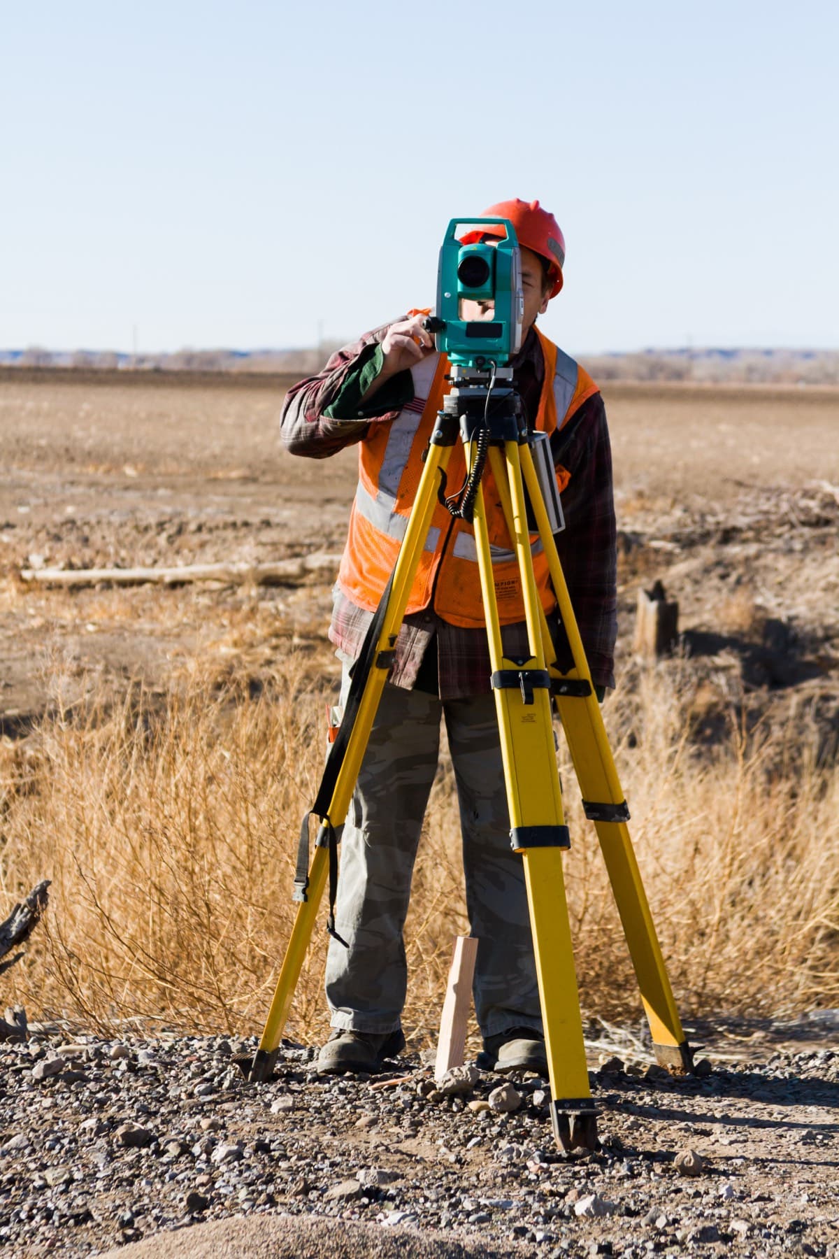 Surveyor in a high-visibility vest sighting through a total station mounted on a yellow tripod on open ground